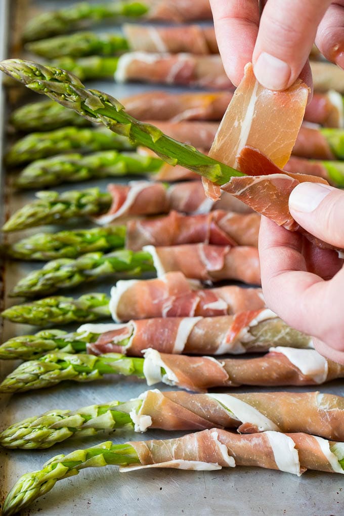 This prosciutto wrapped asparagus is fresh asparagus stalks flavored with savory prosciutto, then broiled to crispy perfection. A hand wrapping a slice of meat around a stalk.