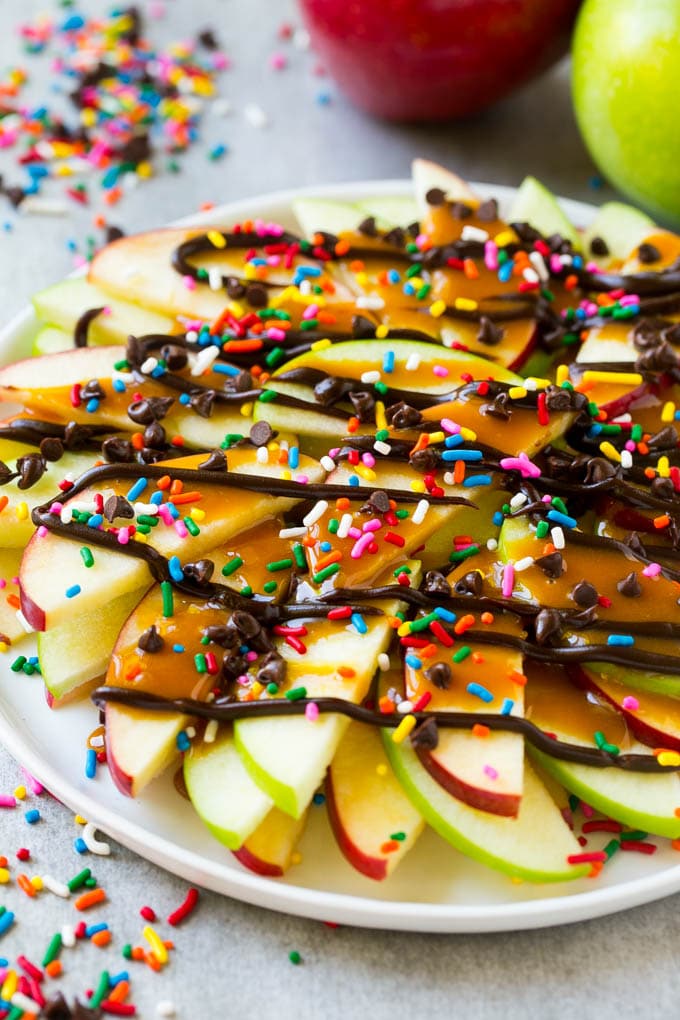 Apple Nachos Recipe | After School Snack | Caramel Apples #apple #apples #caramelapple #snack #dessert #sadud A plate of red and green sliced apples drizzled with caramel and chocolate, then topped with rainbow sprinkles and chocolate chips.