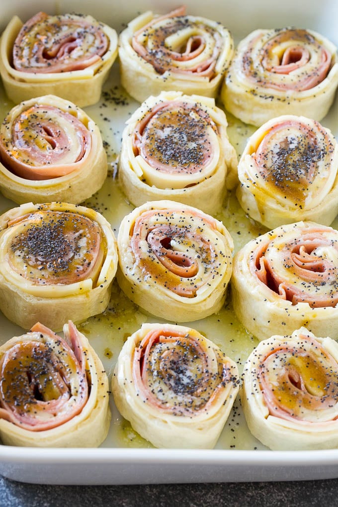 Ham and Cheese Pinwheels Recipe | Ham and Cheese Roll Ups | Ham and Cheese Rolls | Ham and Cheese Bread Rolls #ham #cheese #appetizer #pinwheels #sadud Dough rolled up with ham and cheese and topped with a poppy seed butter in a ceramic dish.