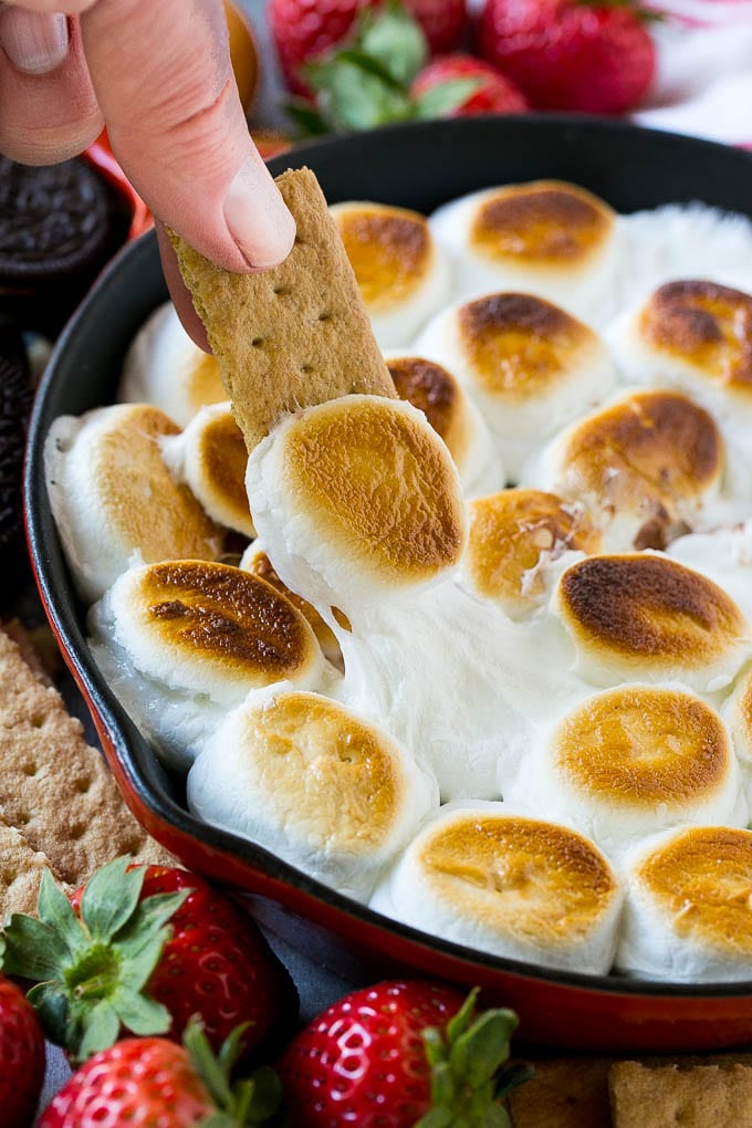 S'mores Dip Recipe | S'mores Skillet | Baked S'mores #smores #dip #chocolate #marshmallow #dessert #sadud A graham cracker topped with melted marshmallow dipping into a skillet of s'mores dip.