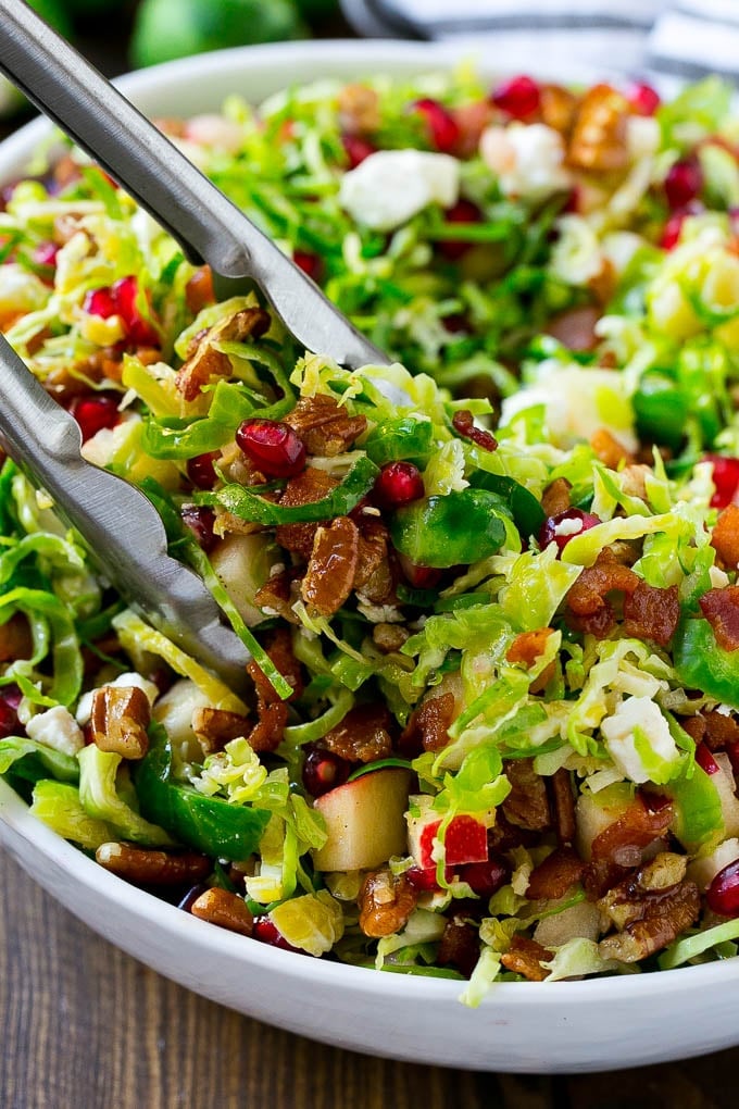 Brussels Sprout Salad Recipe | Shredded Brussels Sprouts Salad | Fall Salad #salad #brusselssprouts #bacon #thanksgiving #pomegranate #apple #dinner #sadud A bowl of brussels sprout salad with tongs picking some up out of the bowl.