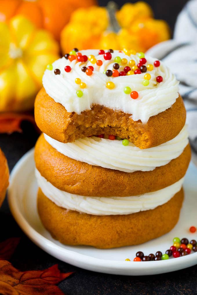 These soft pumpkin cookies bake up light and fluffy, and are topped with a decadent cream cheese frosting and colorful sprinkles. A stack of pumpkin cookies with a bite taken out of one.