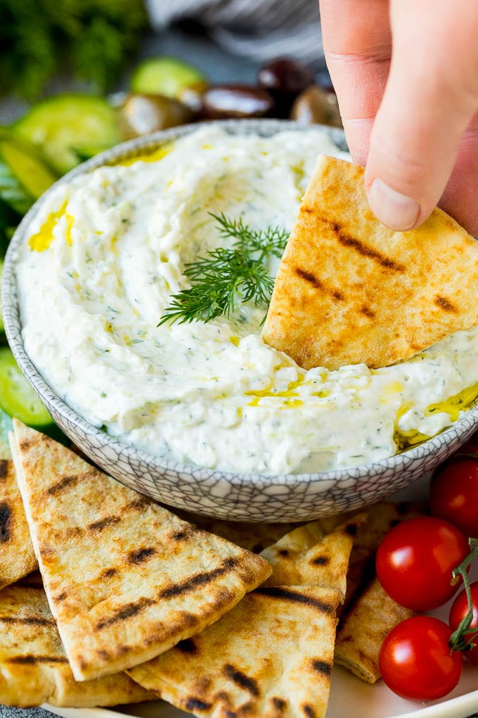 The best homemade tzatziki sauce with cucumber and herbs mixed into Greek yogurt. #appetizer #sadud A hand dipping pita bread into a bowl of tzatziki.