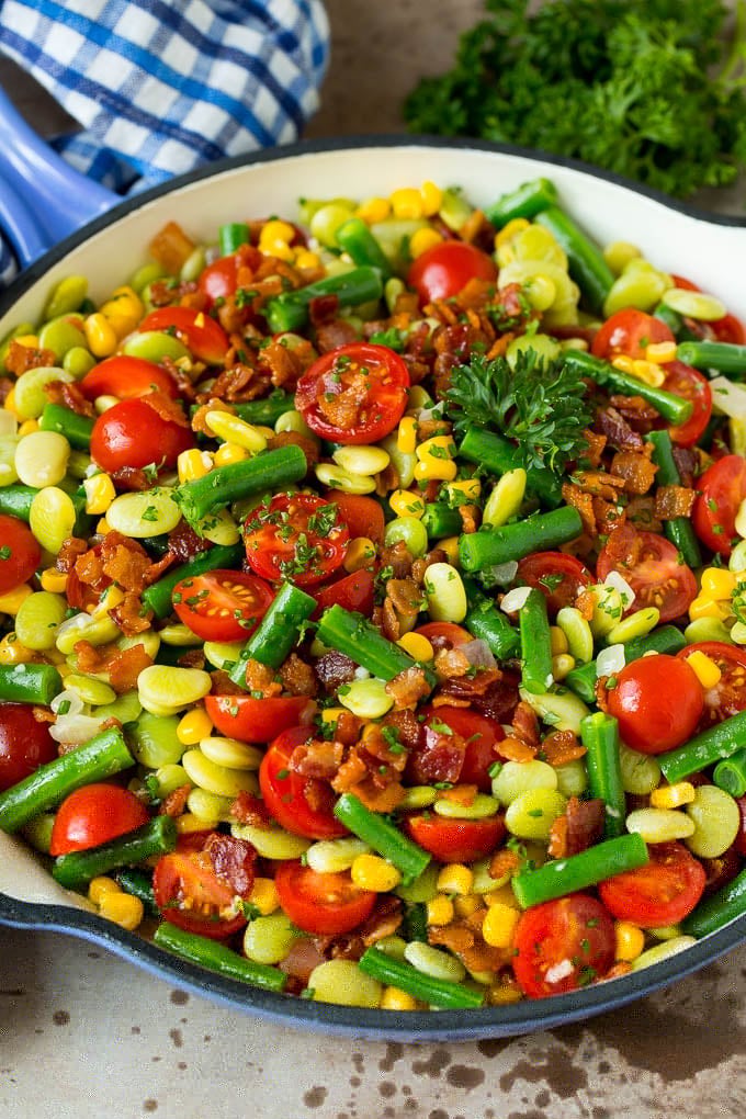 This succotash recipe is a combination of bacon, corn, tomatoes and lima beans, all cooked together to make a colorful and delicious side dish. A skillet of succotash topped with bacon and herbs.