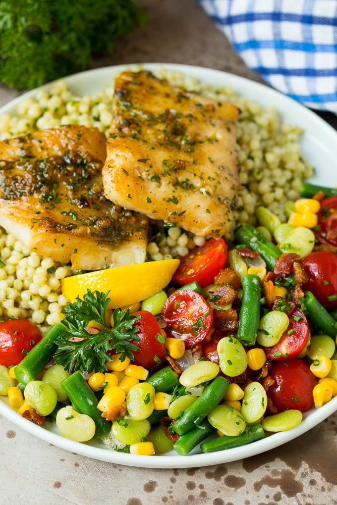 This succotash recipe is a combination of bacon, corn, tomatoes and lima beans, all cooked together to make a colorful and delicious side dish. Succotash served on a plate with fish and couscous.