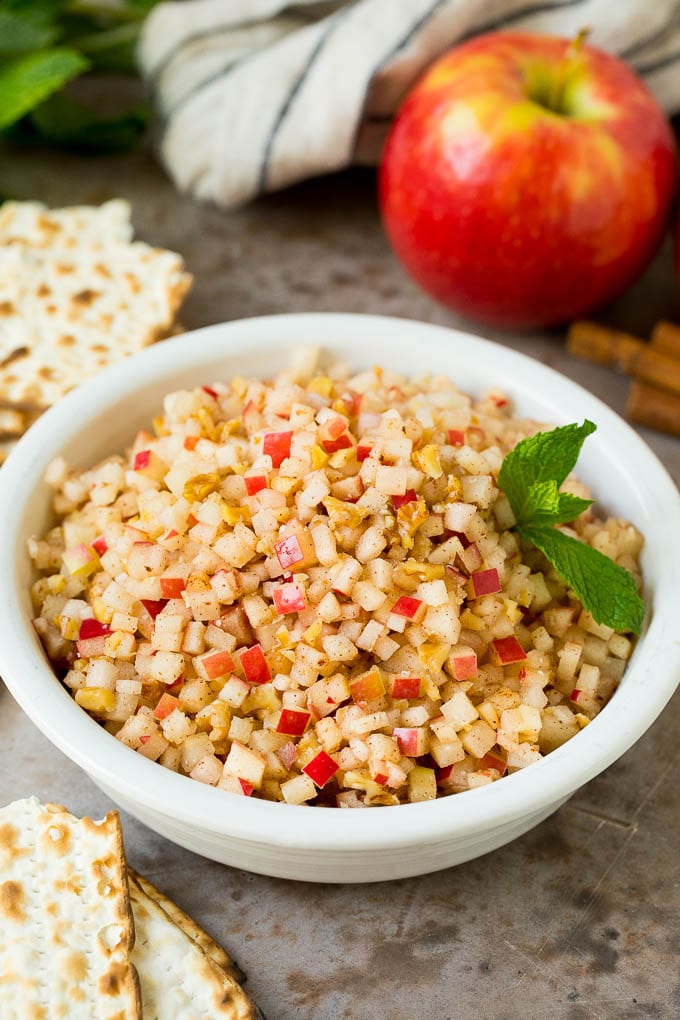 This charoset recipe is a blend of apples, walnuts and honey, all mixed together to make a sweet spread that's an essential part of any Passover meal. A bowl of charoset served with matzo and garnished with mint.