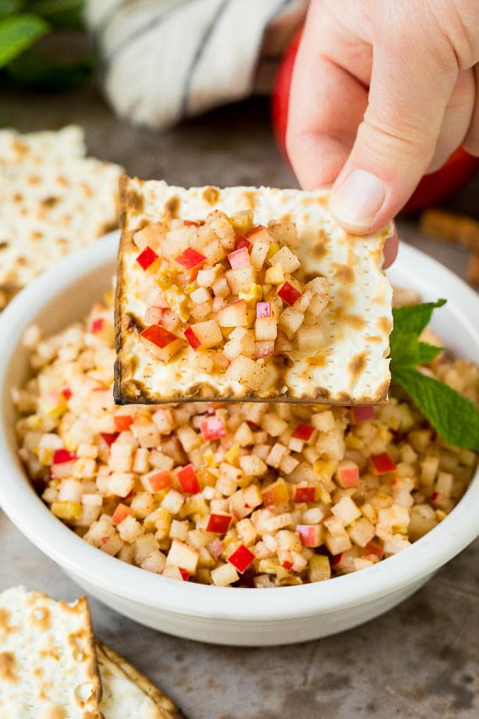 This charoset recipe is a blend of apples, walnuts and honey, all mixed together to make a sweet spread that's an essential part of any Passover meal. A hand holding a piece of matzo with a serving of charoset on top.