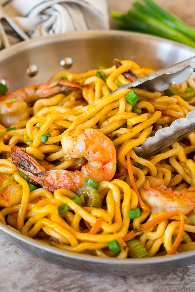 This shrimp lo mein is succulent shrimp with vegetables and egg noodles, all tossed together in a savory sauce. A pan of shrimp lo mein with tongs serving up a portion.