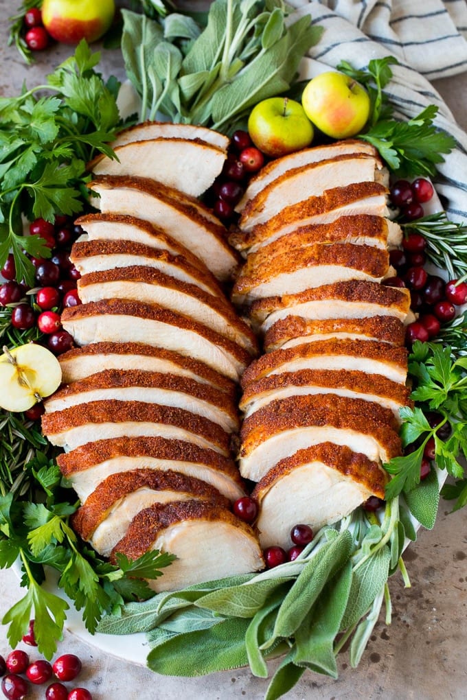 This smoked turkey breast is soaked in brine, then coated in homemade spice rub and cooked on a smoker until it is tender and juicy. Sliced smoked turkey breast on a serving plate with herbs.