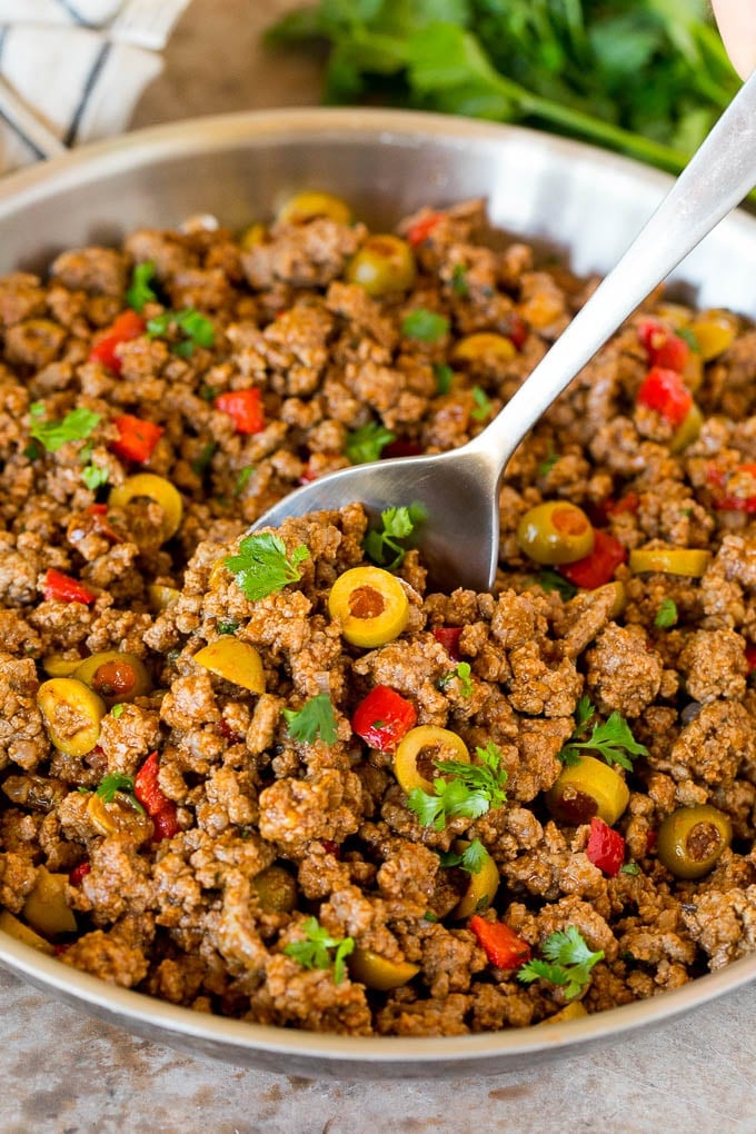 This picadillo is a blend of ground beef, olives and vegetables, all simmered together with spices and tomato sauce. A pan of picadillo with a serving spoon in it.