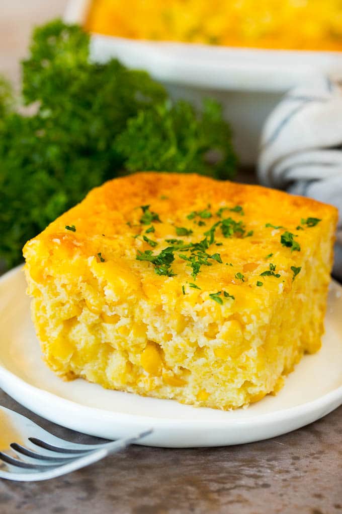 This cornbread casserole is a blend of corn muffin mix, canned corn, creamed corn, seasoning, eggs and butter, all baked together to make a hearty side dish. A square of cornbread casserole topped with parsley.