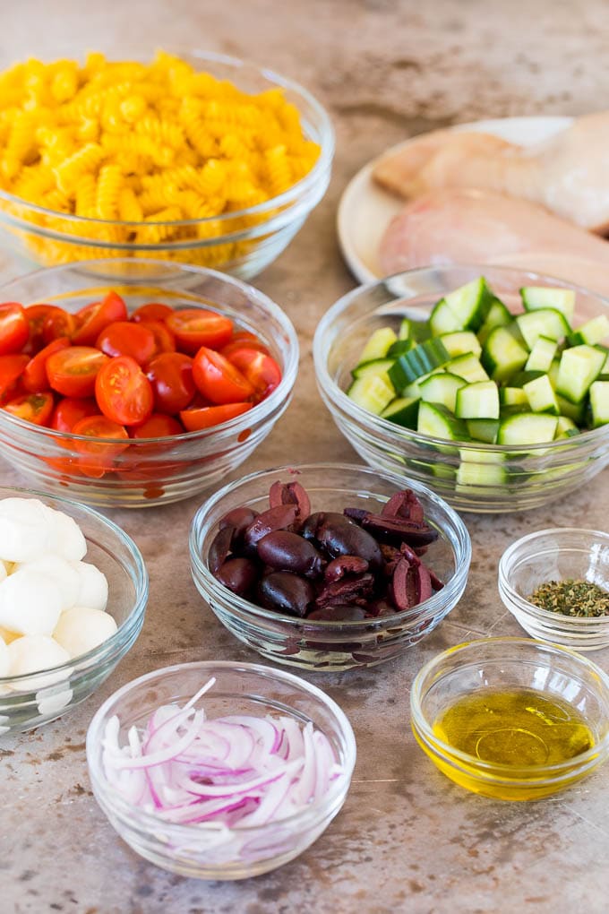 This chicken pasta salad is grilled chicken with olives, cheese and fresh vegetables, all tossed in a homemade Italian dressing. Bowls of vegetables and dry pasta.