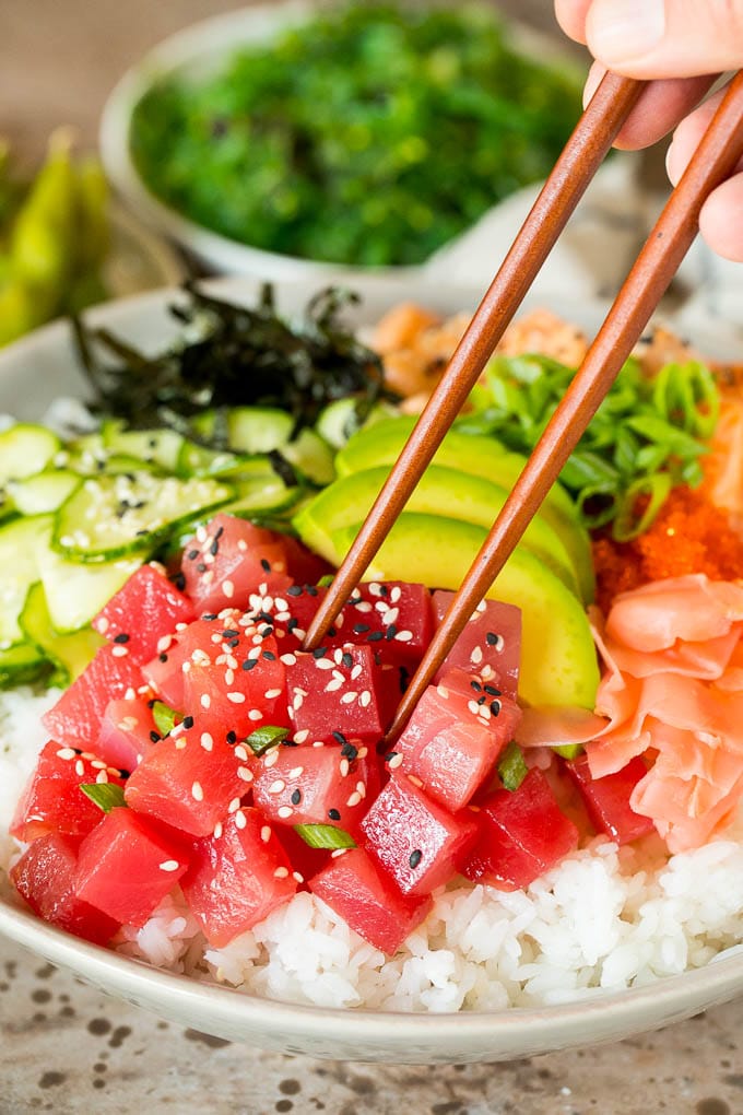 This sushi bowl recipe is assorted fresh fish, avocado, cucumber and seaweed, all served over rice with spicy mayo, picked ginger and tobiko. Chopsticks picking up a piece of ahi tuna in a sushi bowl.