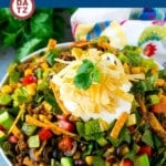 Taco salad in a bowl with ground beef, lettuce, beans, cheese, tomatoes, tortilla strips and avocado, all tossed in a zesty dressing.