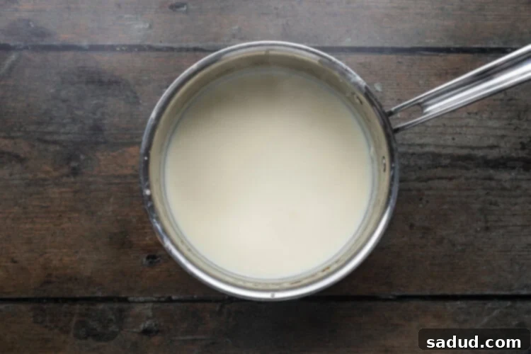 Overhead view of a roux and milk in a silver saucepan on a dark wooden background, being whisked together.