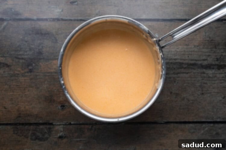 Overhead view of cheese sauce in a silver saucepan on a wooden background, with melted cheese and buffalo sauce.
