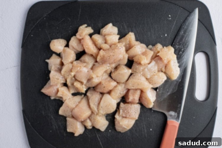 Chopped chicken breasts on a black cutting board with a sharp knife, seasoned with salt and pepper.