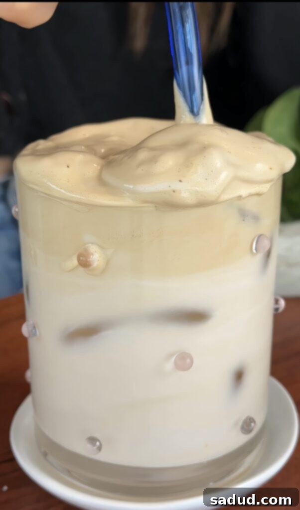 An assembled glass of iced Dalgona whipped honey coffee, showing the distinct layers of milk and the whipped topping.
