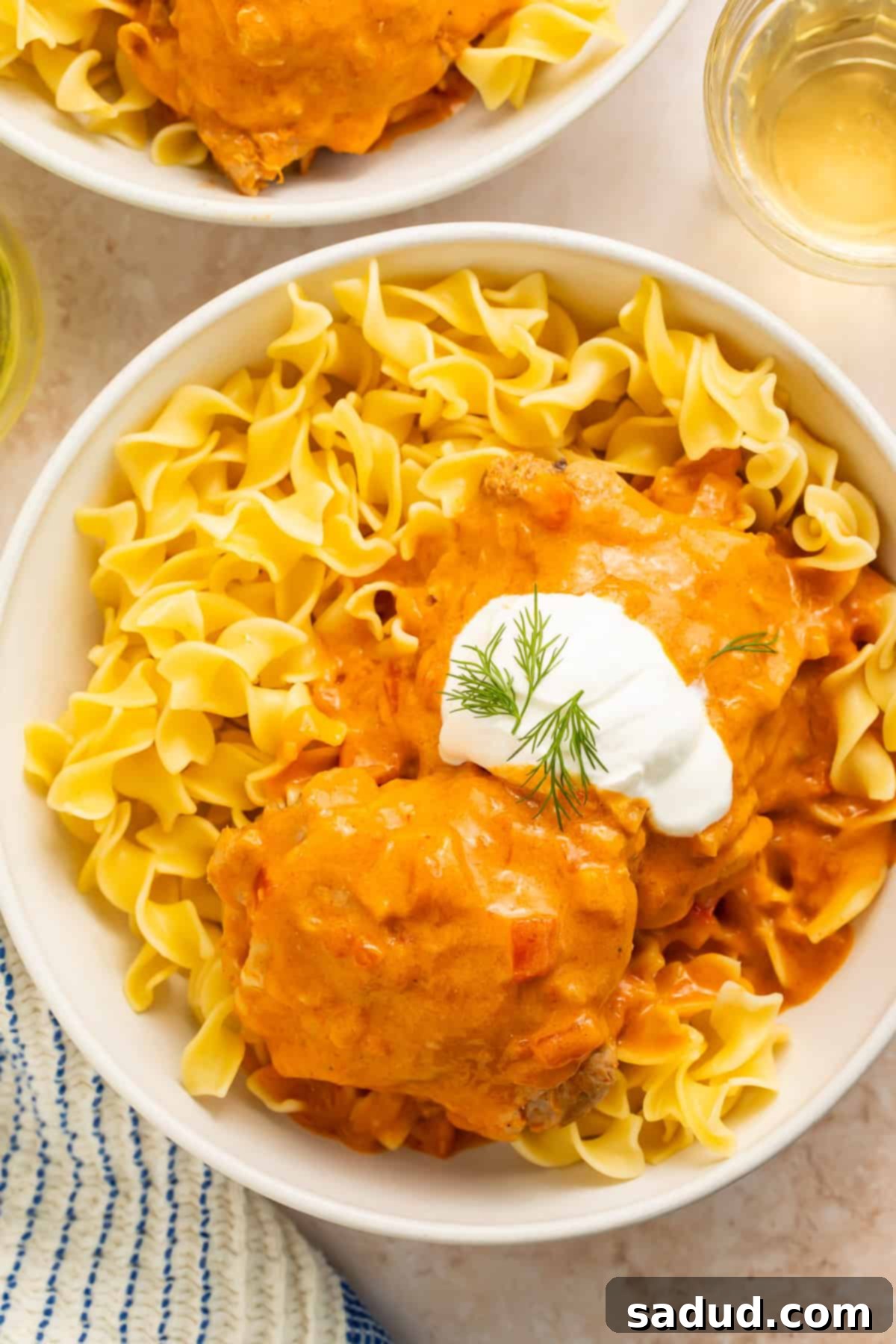 Bowl of creamy tomato and paprika chicken thighs served over egg noodles in a white bowl with a dollop of sour cream on top.