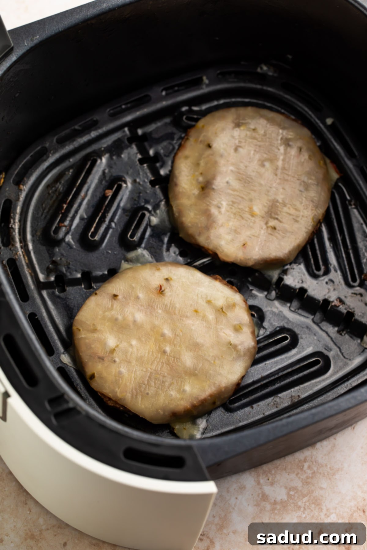 Two frozen burger patties topped with cheese in an air fryer basket.