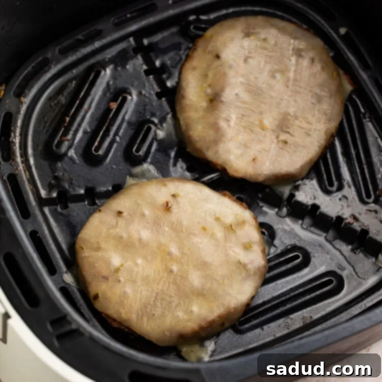 Two frozen burger patties topped with cheese in an air fryer basket.