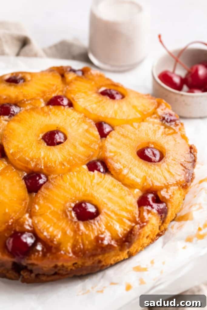 Close-up, 3/4 angle shot of a beautifully baked vegan pineapple upside down cake on a pristine white table, showcasing the golden pineapple and crimson cherries.