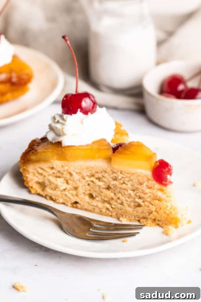 A perfectly cut wedge of vegan pineapple upside down cake, garnished with a dollop of fluffy vegan whipped cream and a bright maraschino cherry, presented on a small dessert plate.
