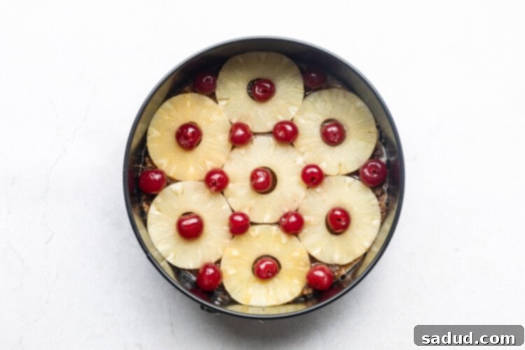Pineapple rings and maraschino cherries in bottom of cake pan on white table.