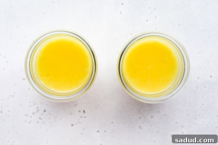 Overhead view of 2 glass jars of bright yellow vegan lemon curd on a white marble background.