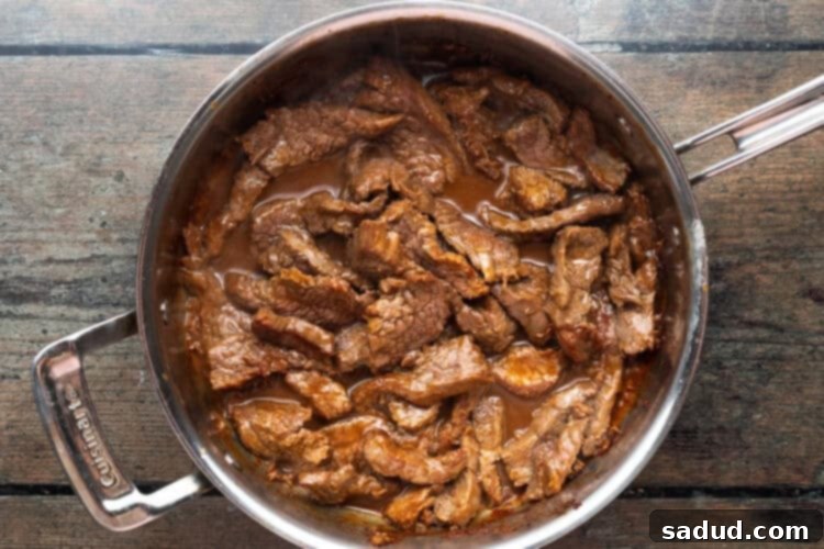 Marinated flank steak searing in a large silver saucepan on a wooden tabletop.
