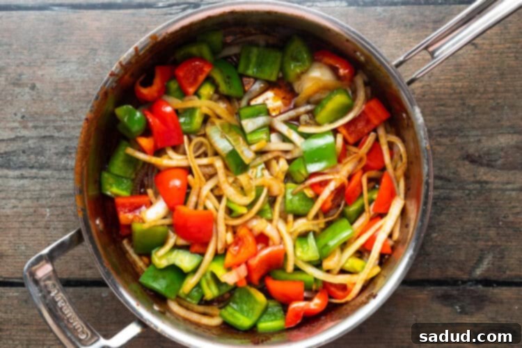 Green and red bell peppers and onions stir-frying in a large silver saucepan on a wooden background.