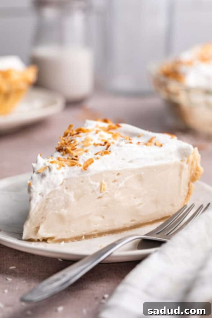 A perfectly sliced wedge of vegan coconut cream pie, topped with a cloud of coconut whipped cream and delicate toasted coconut shavings, resting on a pristine neutral plate beside a fork.
