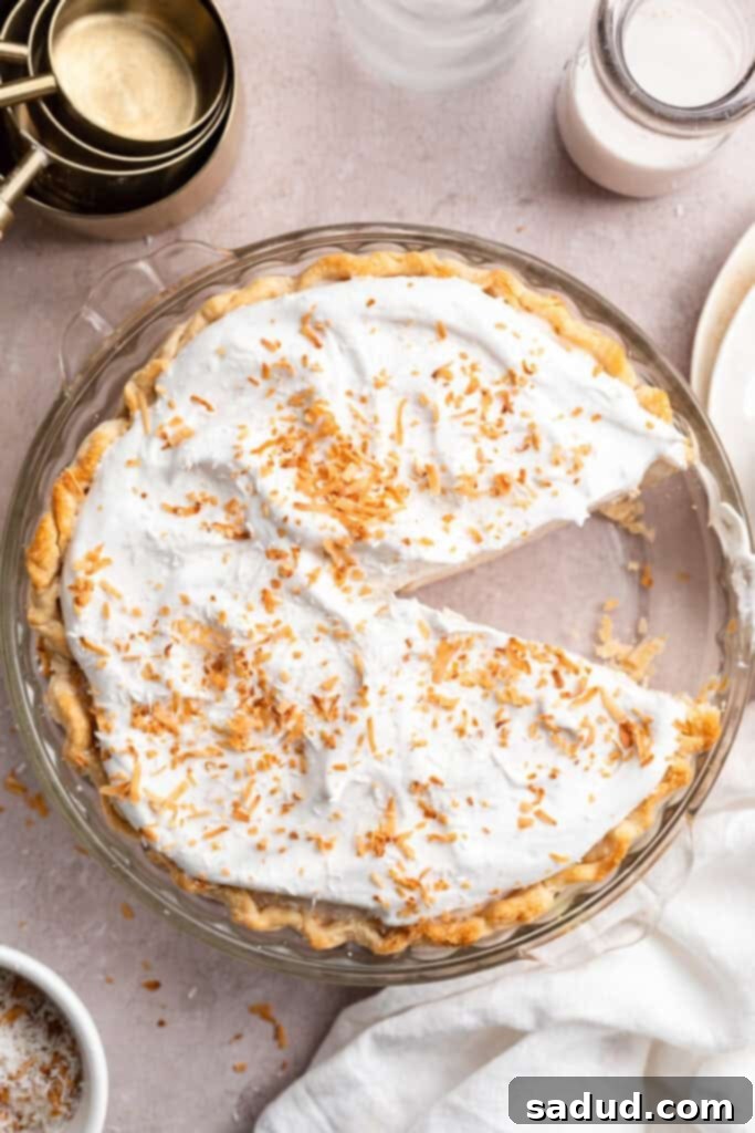 An inviting overhead view of a whole vegan coconut cream pie, beautifully topped with toasted coconut, with one delectable slice already missing, hinting at its irresistible appeal.