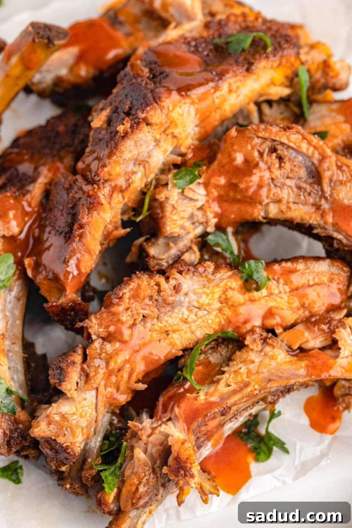 A pile of baked buffalo ribs with bbq sauce on a white plate, garnished with fresh herbs.