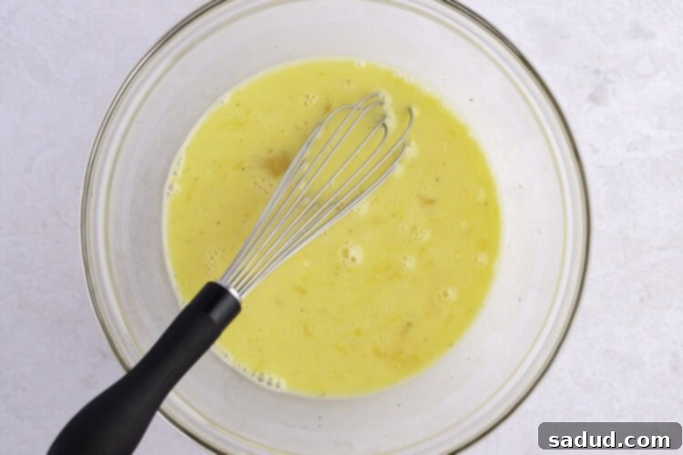 Whisked eggs, milk, salt, and pepper in a large glass mixing bowl with whisk, ready to be cooked.