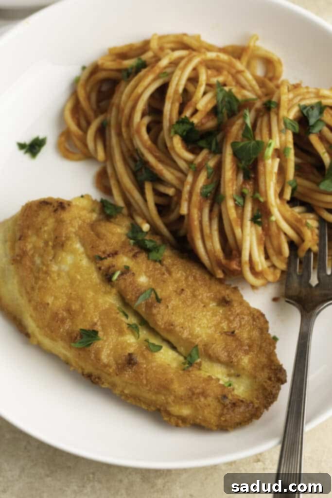 Close-up view of Crusted Chicken Romano served with a side of spaghetti and fresh herbs on a plate.