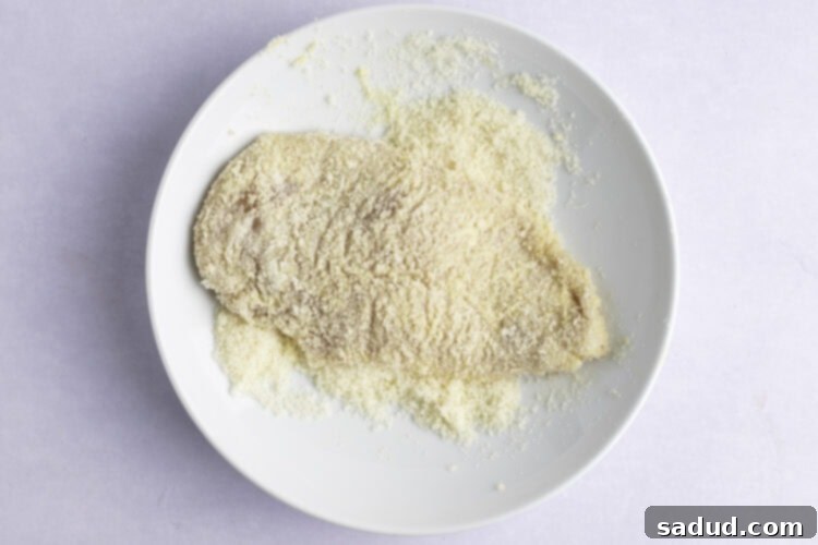 Overhead view of chicken cutlets being dredged in the flour mixture in a large bowl, demonstrating the correct technique for a light, even coating.