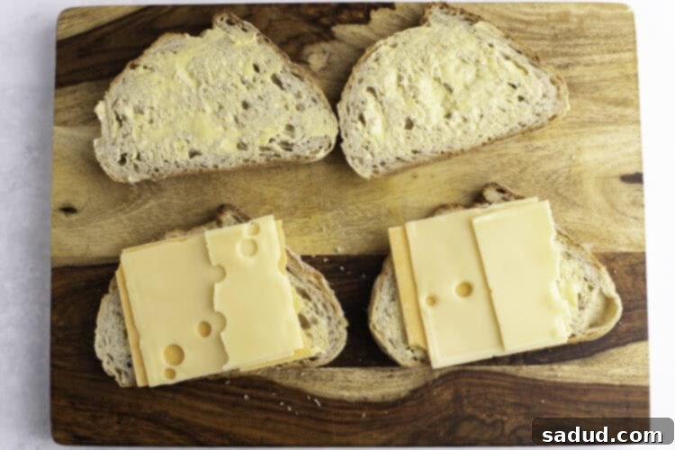 Slices of cheddar cheese and swiss cheese atop slices of buttered sourdough bread on a wooden cutting board.