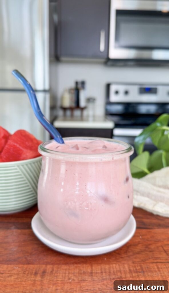 A vibrant pink strawberry watermelon protein yogurt smoothie, garnished with fresh fruit, in a tall glass, set against a bright summer background.