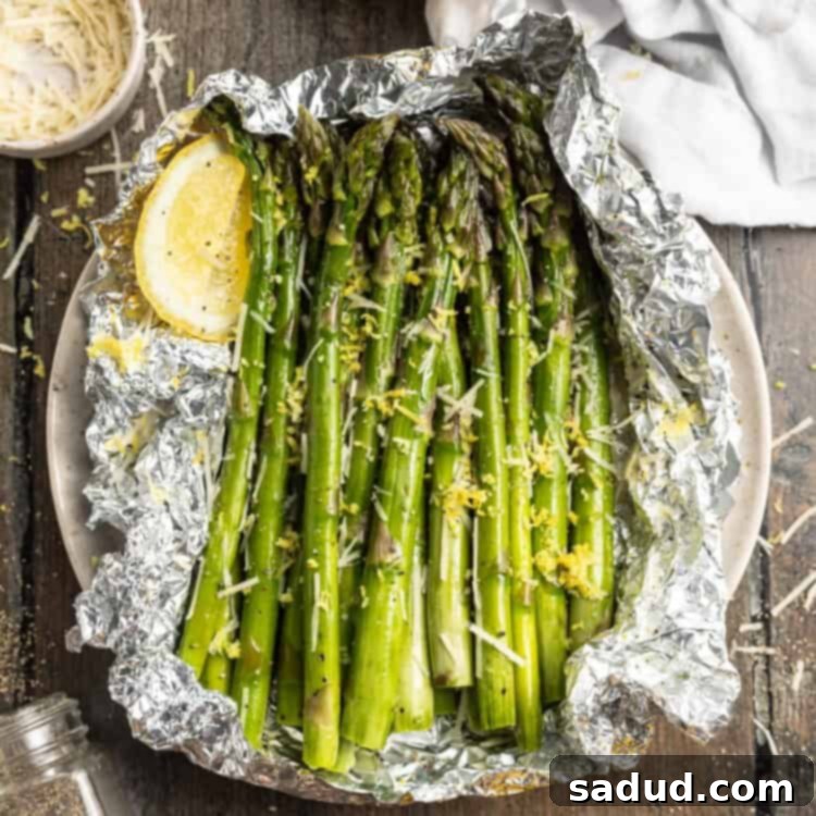 Overhead view of grilled asparagus wrapped in a foil packet resting on a plate with lemon wedges and cheese.