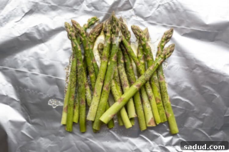 Overhead view of fresh asparagus stalks on a sheet of aluminum foil, seasoned with oil, salt, and pepper.