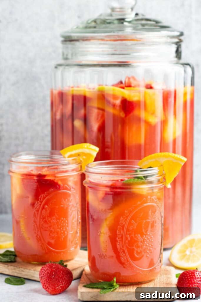 The Ultimate Party Elixir 2 Side view of a large glass container of bright red jungle juice behind 2 small mason jars of jungle juice garnished with fruit and a clear straw.