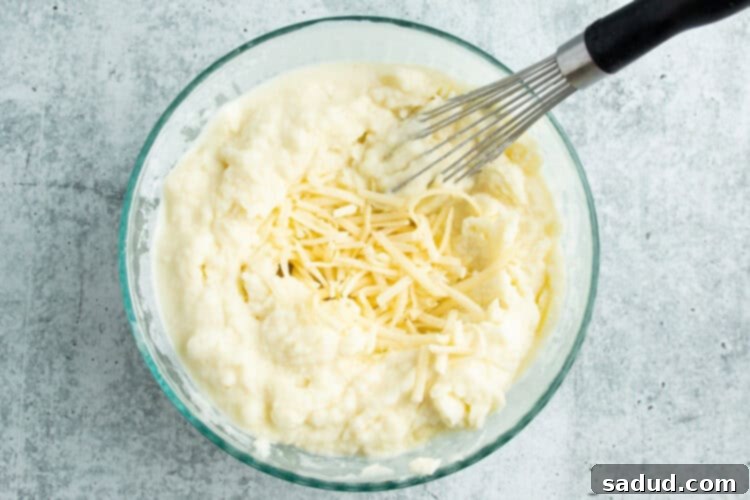 Overhead view of creamy potato mixture in a large bowl with shredded white cheese and a whisk, ready for folding.