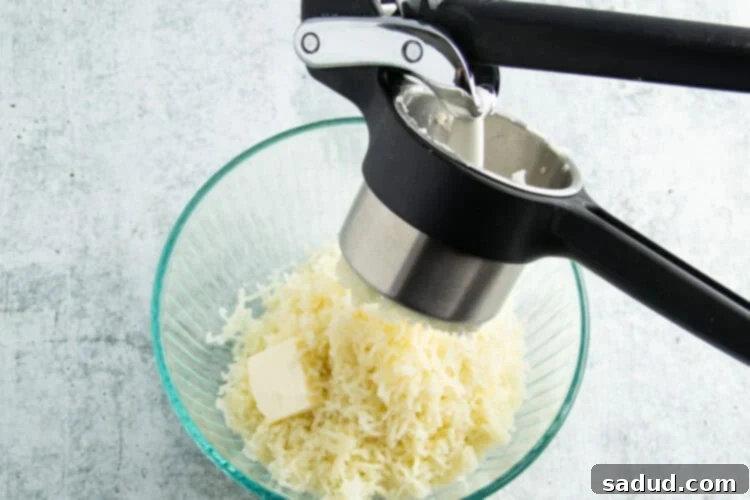 Potatoes being pressed through potato ricer and into a large glass mixing bowl, creating a light, fluffy texture.