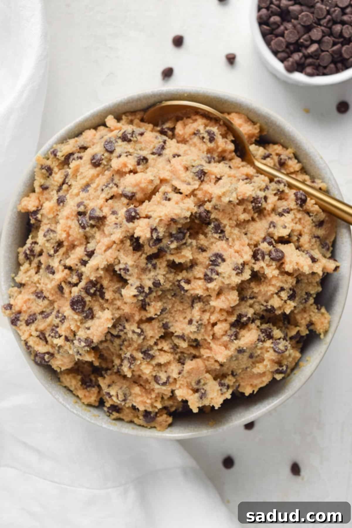 Overhead view of a bowl of creamy keto edible chocolate chip cookie dough with a gold spoon, set on a bright white tabletop. Scattered chocolate chips add to the visual appeal.