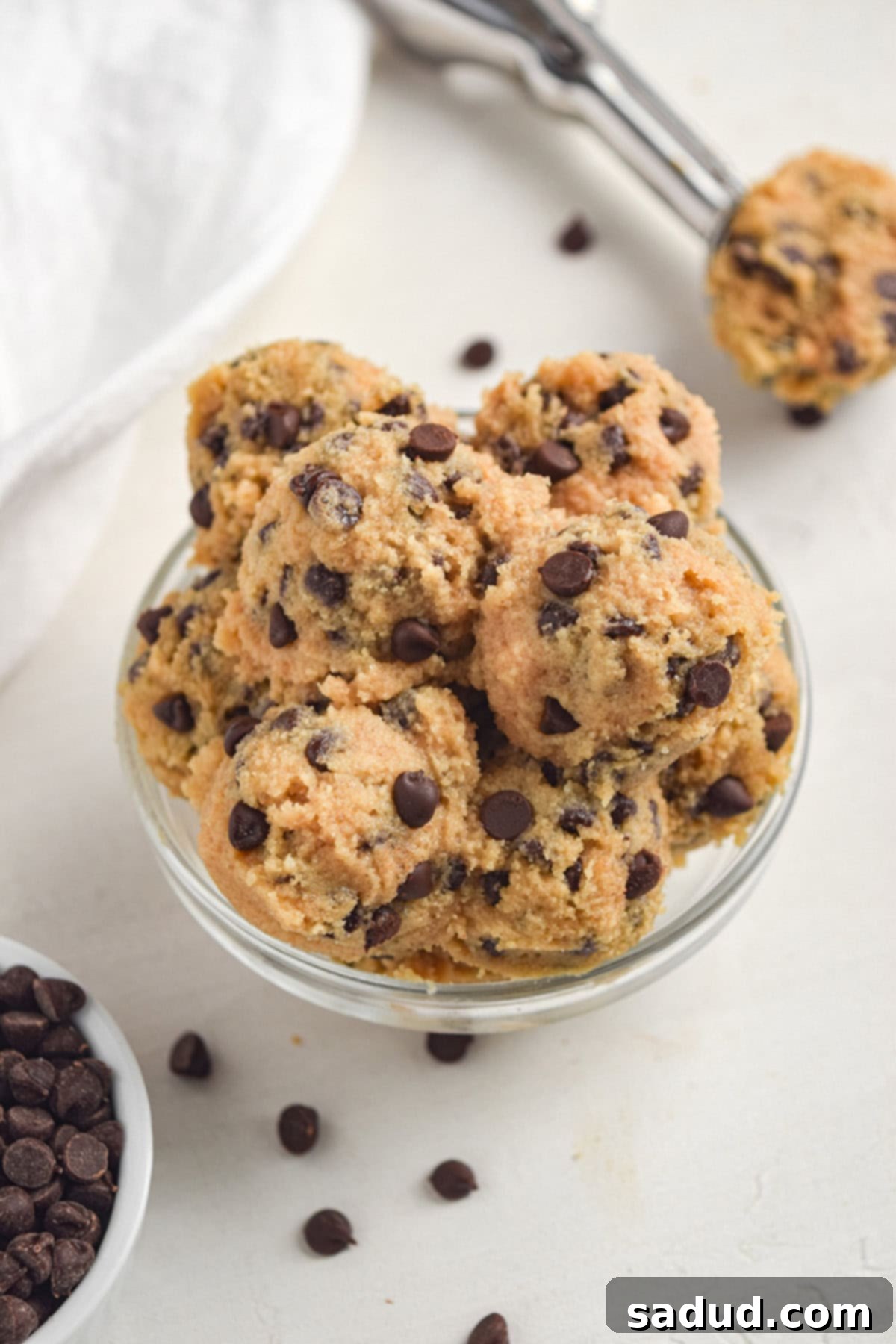 A clear glass mixing bowl filled with scoops of ready-to-eat keto chocolate chip cookie dough, accompanied by scattered sugar-free chocolate chips on a white tabletop.