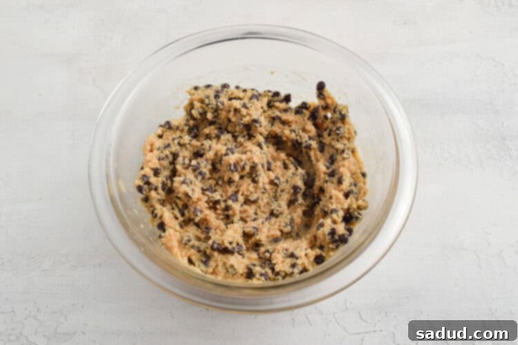 Overhead view of keto chocolate chip cookie dough in a small glass mixing bowl on a white table, after chocolate chips have been folded in.