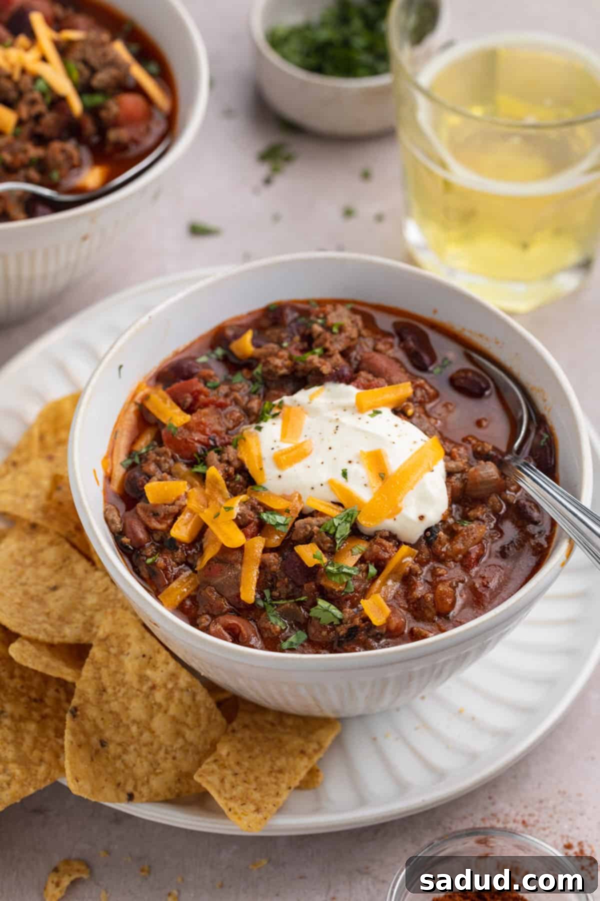 Angled view of a white bowl holding deep red Mexican chili topped with sour cream and shredded cheddar cheese next to tortilla chips on a white plate, with fresh cilantro garnish.
