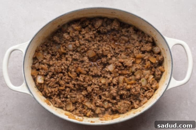 Overhead view of seasoned, crumbled ground beef in a large chili pot with tender chopped onion and minced garlic on a white table, ready for simmering.