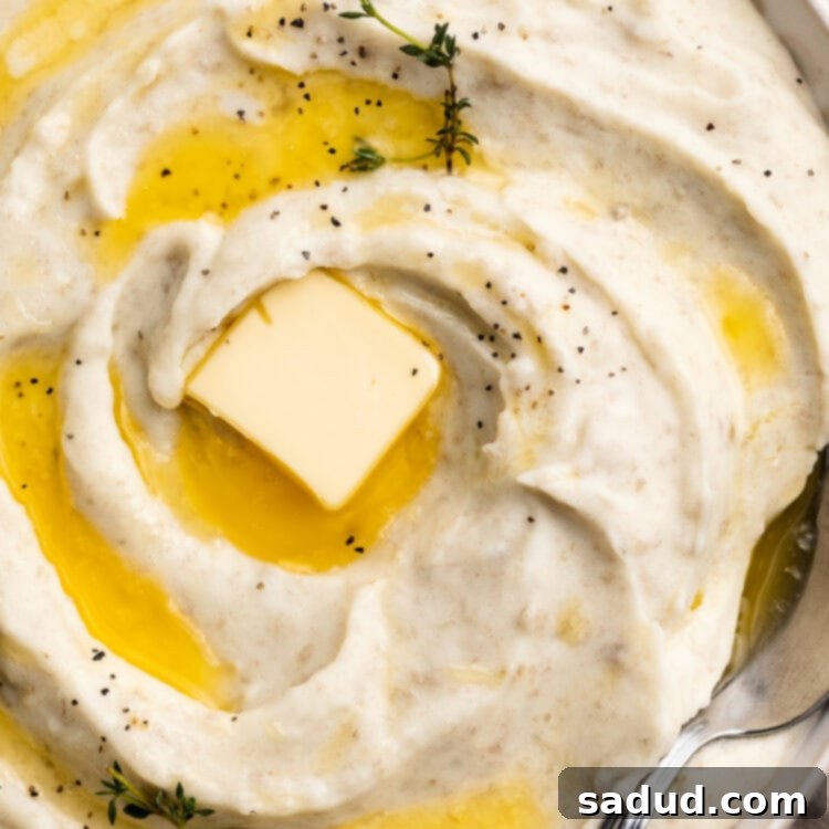 Close-up, overhead view of a large bowl of creamy whipped potatoes, swirled with melted butter and topped with a pat of butter and fresh herbs.